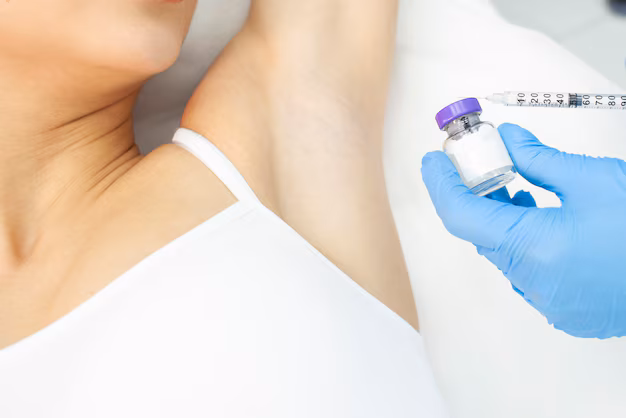 A close-up shot of a doctor's hand in a blue glove holding a small vial labeled "Botox" and a syringe, ready to inject a woman's armpit. The woman's white tank top is visible. The image emphasizes the product and the procedure.