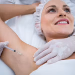 A doctor in a white coat and gloves is injecting a woman's armpit with a syringe. The woman, wearing a hair net and with her eyes closed, appears relaxed. The focus is on the injection process.