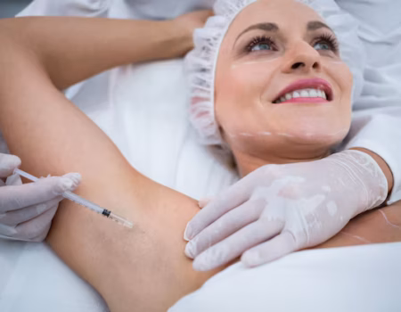 A doctor in a white coat and gloves is injecting a woman's armpit with a syringe. The woman, wearing a hair net and with her eyes closed, appears relaxed. The focus is on the injection process.