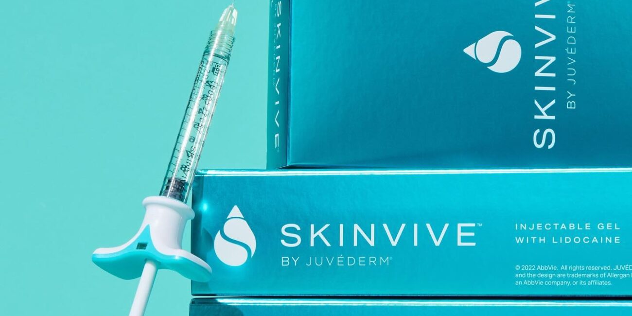 A product shot showing a single SKINVIVE by JUVÉDERM injectable gel syringe leaning against a stack of three product boxes. The packaging is a vibrant blue, and the text on the box identifies it as "INJECTABLE GEL with LIDOCAINE." The image highlights the product's branding and form.
