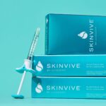 A product shot showing a single SKINVIVE by JUVÉDERM injectable gel syringe leaning against a stack of three product boxes. The packaging is a vibrant blue, and the text on the box identifies it as "INJECTABLE GEL with LIDOCAINE." The image highlights the product's branding and form.