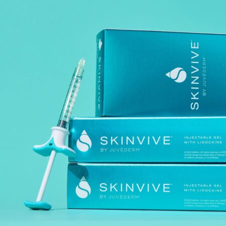 A product shot showing a single SKINVIVE by JUVÉDERM injectable gel syringe leaning against a stack of three product boxes. The packaging is a vibrant blue, and the text on the box identifies it as "INJECTABLE GEL with LIDOCAINE." The image highlights the product's branding and form.