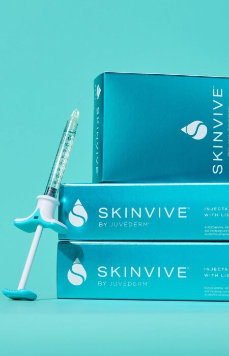 A product shot showing a single SKINVIVE by JUVÉDERM injectable gel syringe leaning against a stack of three product boxes. The packaging is a vibrant blue, and the text on the box identifies it as "INJECTABLE GEL with LIDOCAINE." The image highlights the product's branding and form.