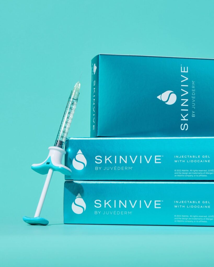 A product shot showing a single SKINVIVE by JUVÉDERM injectable gel syringe leaning against a stack of three product boxes. The packaging is a vibrant blue, and the text on the box identifies it as "INJECTABLE GEL with LIDOCAINE." The image highlights the product's branding and form.