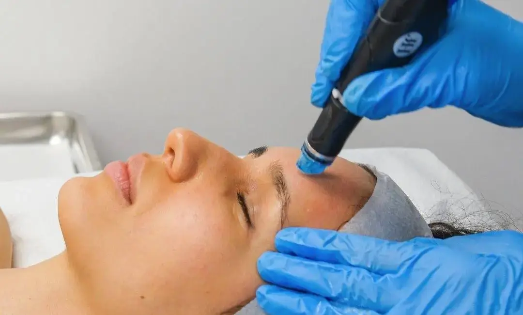 An esthetician performing a HydraFacial treatment on a client in a modern Delhi clinic. The HydraFacial machine is visible in the background, showing the vortex-fusion technology being used.