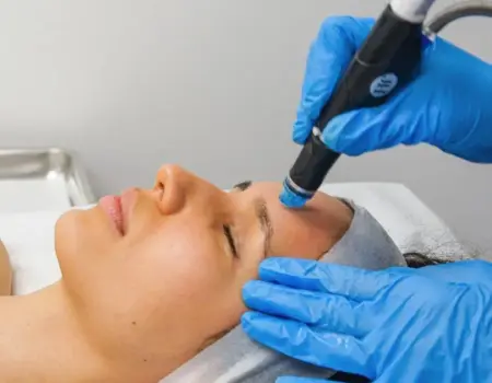 An esthetician performing a HydraFacial treatment on a client in a modern Delhi clinic. The HydraFacial machine is visible in the background, showing the vortex-fusion technology being used.