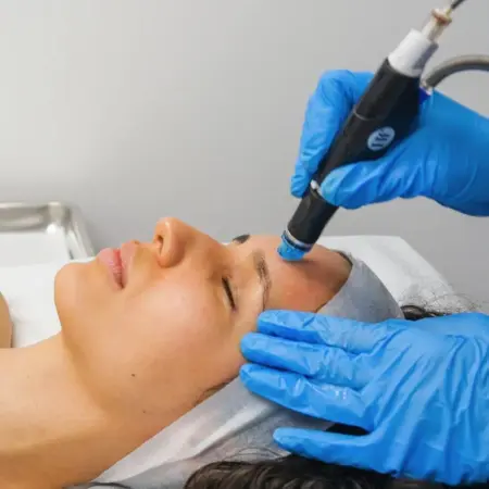 An esthetician performing a HydraFacial treatment on a client in a modern Delhi clinic. The HydraFacial machine is visible in the background, showing the vortex-fusion technology being used.