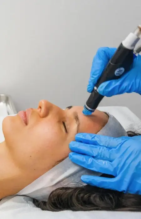 An esthetician performing a HydraFacial treatment on a client in a modern Delhi clinic. The HydraFacial machine is visible in the background, showing the vortex-fusion technology being used.